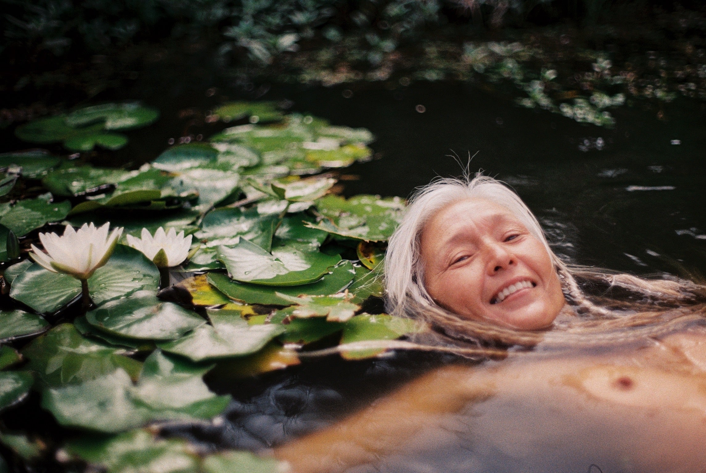 An elder woman with glowing skin and silver braided hair floats on her back in a lake amidst lily pads and lotuses, glancing up with a relaxed smile